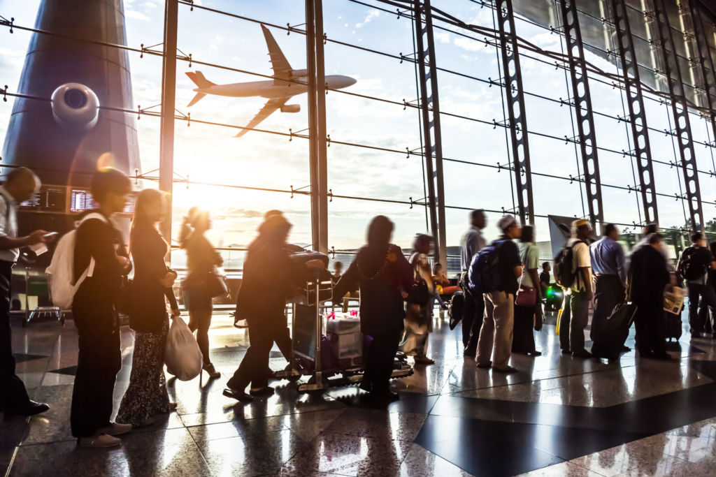 Ein Flugzeug startet an einem Flughafen, im Vordegrund warten Reisende am Check-In Schalter.